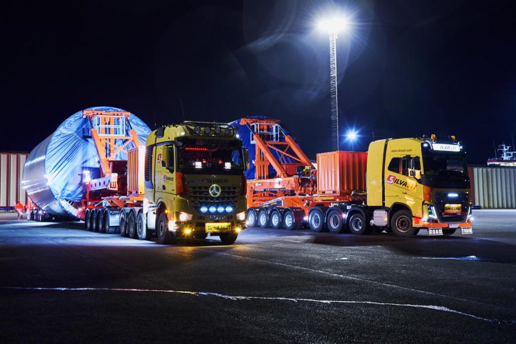 Two yellow Silvasti trucks with red clamp trailers that have wind turbine tower sections loaded in them.